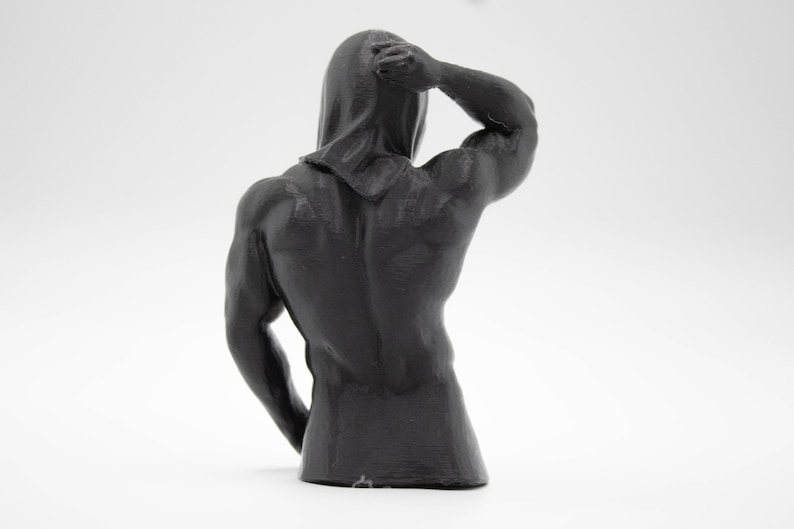Puede incluir: Escultura gris oscuro de una figura musculosa, vista desde atr&aacute;s. La figura lleva una capucha y tiene un brazo levantado, tocando la cabeza. La escultura est&aacute; sobre un fondo blanco.