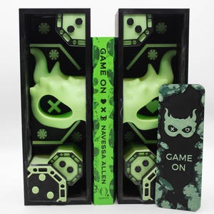 May include: Black bookends with neon green accents and a book titled "Game On" by Navessa Allen. The bookends feature a mask design and geometric shapes. A matching bookmark with the text "Game On" is also present.