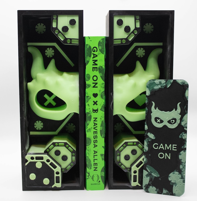 May include: Black bookends with neon green accents and a book titled "Game On" by Navessa Allen. The bookends feature a mask design and geometric shapes. A matching bookmark with the text "Game On" is also present.