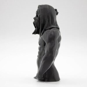 Puede incluir: Busto gris oscuro de una figura con cabeza encapuchada y torso musculoso. La figura tiene una textura detallada y parece ser una escultura o figura. El fondo es blanco liso, enfatizando la forma y el detalle del objeto.