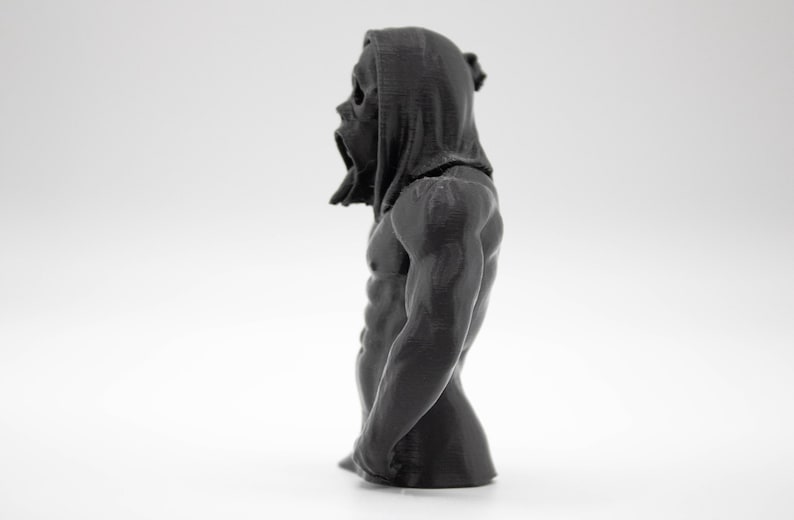 Puede incluir: Busto gris oscuro de una figura con cabeza encapuchada y torso musculoso. La figura tiene una textura detallada y parece ser una escultura o figura. El fondo es blanco liso, enfatizando la forma y el detalle del objeto.