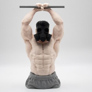May include: A muscular torso figurine wearing a black gas mask and grey shorts, depicted with arms raised overhead while gripping a horizontal grey bar. The sculpture features detailed skin texture and defined abdominal muscles in a light skin tone.