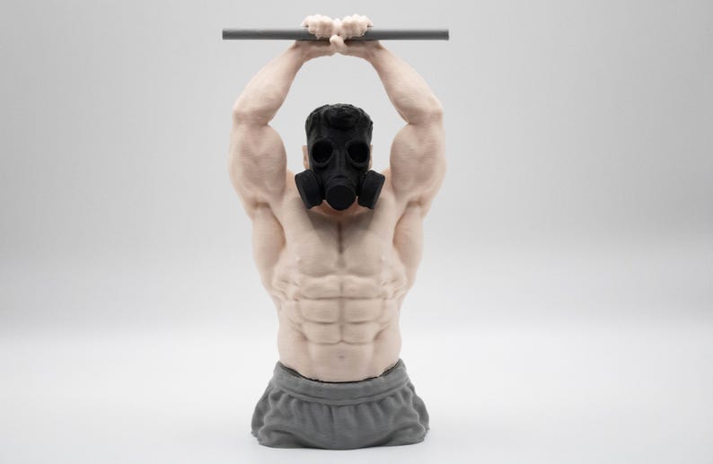 May include: A muscular torso figurine wearing a black gas mask and grey shorts, depicted with arms raised overhead while gripping a horizontal grey bar. The sculpture features detailed skin texture and defined abdominal muscles in a light skin tone.