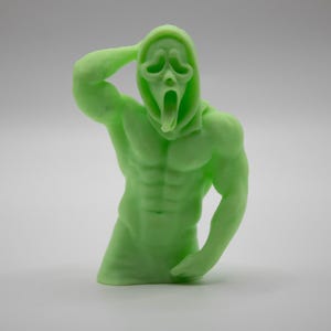 Puede incluir: Una figura musculosa verde claro del personaje Ghostface de la serie de pel&iacute;culas de terror Scream. La figura est&aacute; posada con un brazo levantado hacia la cabeza y el otro brazo doblado por el codo. La figura tiene una capucha y una m&aacute;scara gritando.