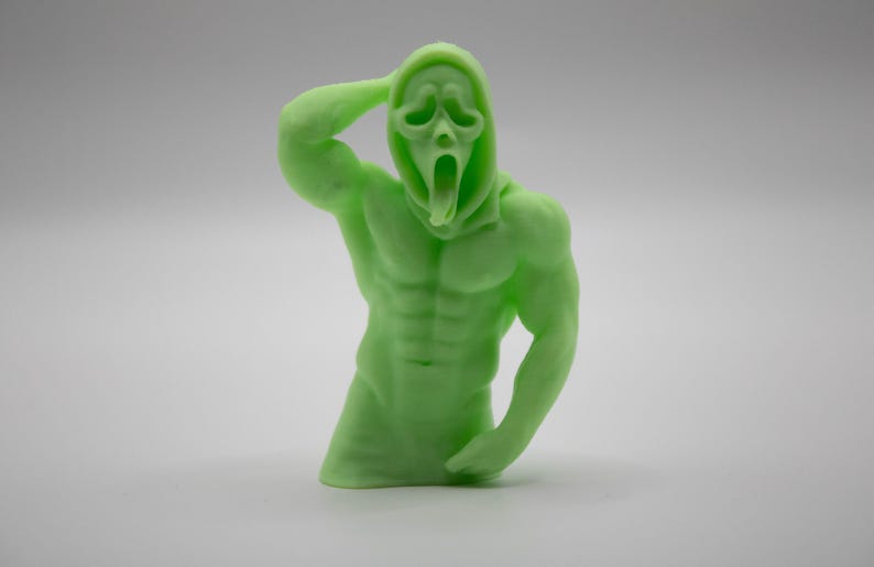 Puede incluir: Una figura musculosa verde claro del personaje Ghostface de la serie de pel&iacute;culas de terror Scream. La figura est&aacute; posada con un brazo levantado hacia la cabeza y el otro brazo doblado por el codo. La figura tiene una capucha y una m&aacute;scara gritando.