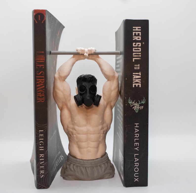 May include: A bookend featuring a muscular torso figure wearing a black gas mask, positioned between two books titled Little Stranger by Leigh Rivers and Her Soul to Take by Harley Laroux, with the figure gripping a horizontal metal bar overhead.