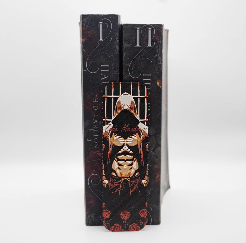 May include: A black bookmark featuring a hooded figure with a muscular torso, set against a prison bar design. The bookmark is placed between two books with dark covers and the text "II" and "I". Red roses adorn the bottom of the bookmark.