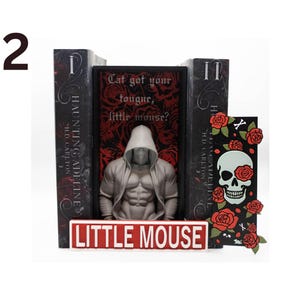 May include: A bookend set featuring a muscular figure in a hooded jacket, flanked by two books and a decorative card. The books are titled "Haunting Adeline" and the card has a skull with roses. The set includes a sign that reads "Little Mouse".