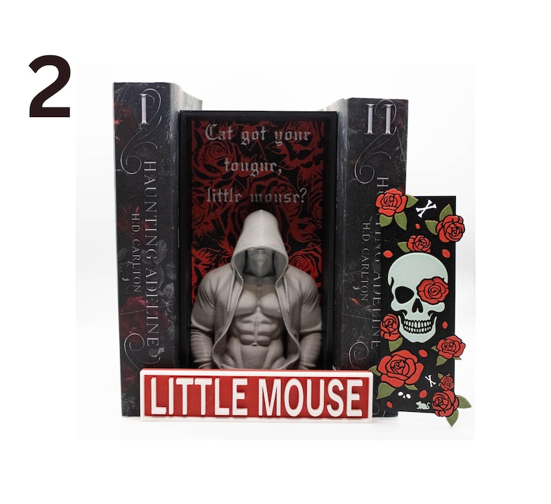 May include: A bookend set featuring a muscular figure in a hooded jacket, flanked by two books and a decorative card. The books are titled "Haunting Adeline" and the card has a skull with roses. The set includes a sign that reads "Little Mouse".
