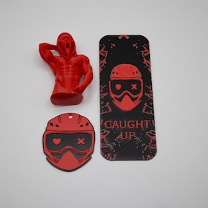 Pode incluir: Uma estatueta vermelha de uma pessoa com a m&atilde;o na cabe&ccedil;a, um porta-chaves vermelho em forma de capacete e uma etiqueta retangular preta e vermelha com as palavras "CAUGHT UP". A etiqueta e o porta-chaves apresentam um design de capacete.