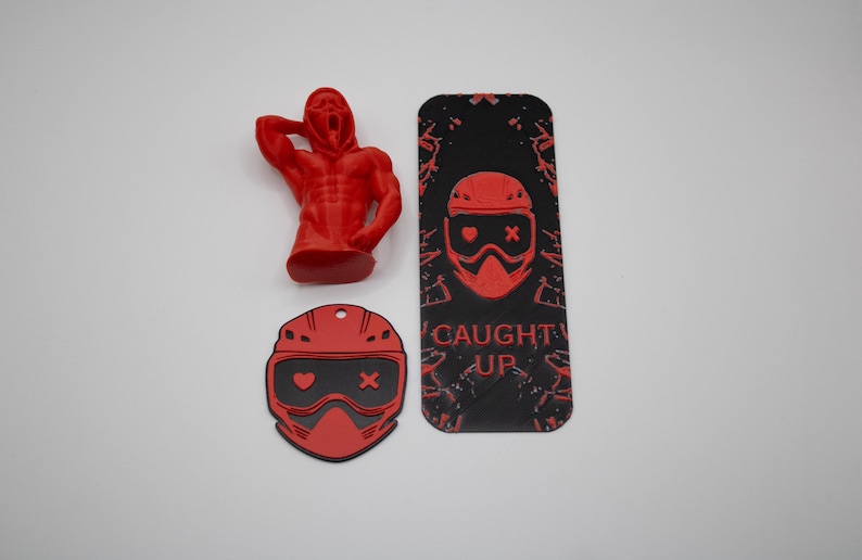 Pode incluir: Uma estatueta vermelha de uma pessoa com a m&atilde;o na cabe&ccedil;a, um porta-chaves vermelho em forma de capacete e uma etiqueta retangular preta e vermelha com as palavras "CAUGHT UP". A etiqueta e o porta-chaves apresentam um design de capacete.