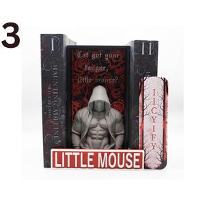 May include: A bookend set featuring a muscular figure in a hooded sweatshirt, flanked by two books and a decorative piece. The central book has the text "Cat got your tougur, little mouse?" and the base reads "LITTLE MOUSE".