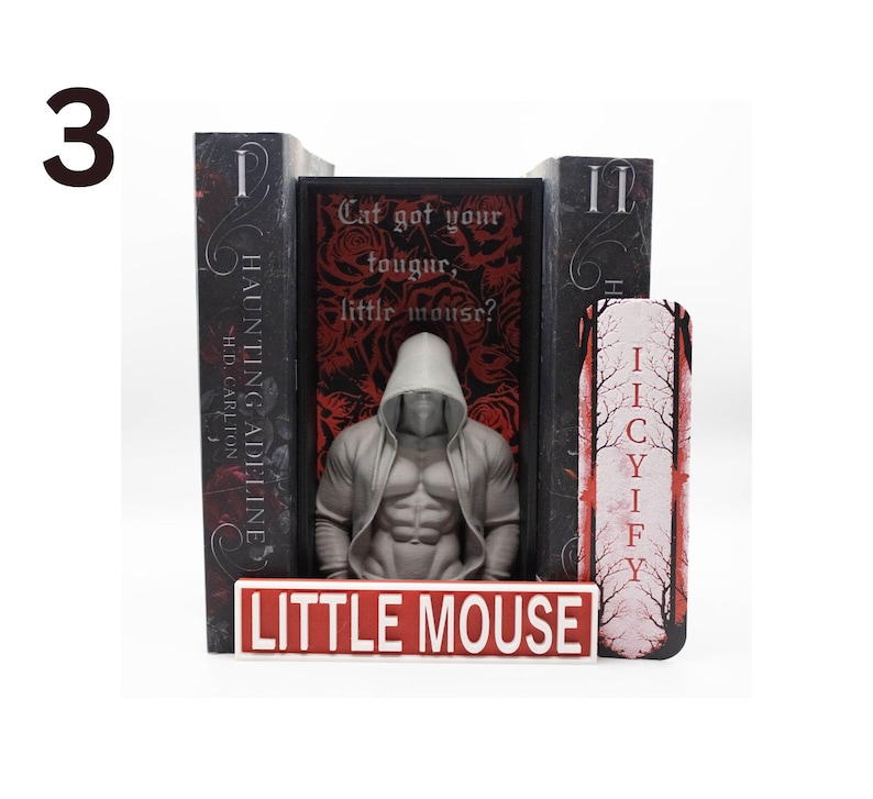 May include: A bookend set featuring a muscular figure in a hooded sweatshirt, flanked by two books and a decorative piece. The central book has the text "Cat got your tougur, little mouse?" and the base reads "LITTLE MOUSE".