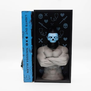 May include: A book titled "LIGHTS OUT" by Navessa Allen stands next to a gray sculpture of a muscular figure wearing a blue skull mask. The sculpture is inside a black box decorated with blue hearts, stethoscopes, and skulls. The book has a blue cover.