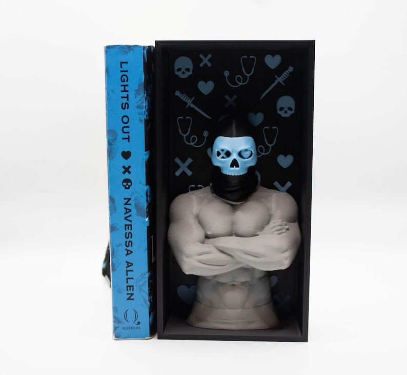 May include: A book titled "LIGHTS OUT" by Navessa Allen stands next to a gray sculpture of a muscular figure wearing a blue skull mask. The sculpture is inside a black box decorated with blue hearts, stethoscopes, and skulls. The book has a blue cover.