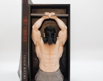 Puede incluir: Un sujetalibros que muestra una figura musculosa con una máscara de gas negra, con los brazos levantados y las manos agarradas a una barra horizontal. La figura está colocada dentro de un marco rectangular negro junto al libro Little Stranger de Leigh Rivers.