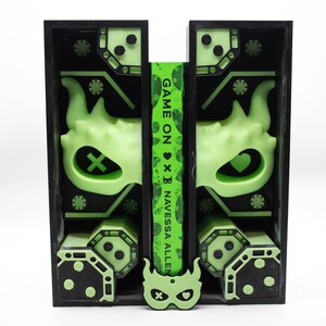 May include: A black and green art piece with a neon green design. The design includes a mask, geometric shapes, and the text "GAME ON x B NAVESSA ALLEN" on a green strip. The piece has a symmetrical layout and a modern aesthetic.