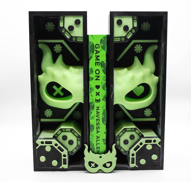 May include: A black and green art piece with a neon green design. The design includes a mask, geometric shapes, and the text "GAME ON x B NAVESSA ALLEN" on a green strip. The piece has a symmetrical layout and a modern aesthetic.