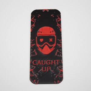 Pode incluir: Marcador de livros retangular preto com um gr&aacute;fico vermelho de um rosto com capacete e &oacute;culos e as palavras "CAUGHT UP". O design tem um efeito de respingo.