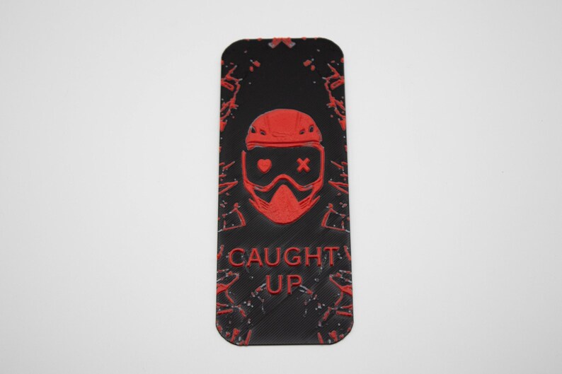 Pode incluir: Marcador de livros retangular preto com um gr&aacute;fico vermelho de um rosto com capacete e &oacute;culos e as palavras "CAUGHT UP". O design tem um efeito de respingo.