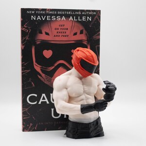 May include: A book titled Caught Up by Navessa Allen featuring a red helmet graphic and the text Get On Your Knees And Prey, paired with a muscular torso figurine wearing a red helmet and black gloves, positioned in front of the book cover.