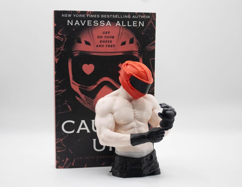 May include: A book titled Caught Up by Navessa Allen featuring a red helmet graphic and the text Get On Your Knees And Prey, paired with a muscular torso figurine wearing a red helmet and black gloves, positioned in front of the book cover.