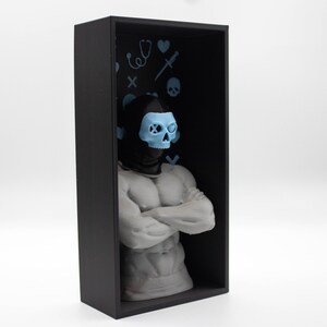 May include: A black rectangular display case houses a white muscular torso sculpture with arms crossed. The sculpture wears a black hood and a light blue skull mask. The case's black interior features light blue medical-themed icons.