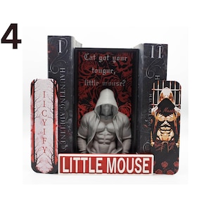 May include: A book display featuring a muscular, hooded figure statue, flanked by decorative bookends. The bookends have text that reads "IICYIFY" and "HAUNTING ADELINE". A sign at the base reads "LITTLE MOUSE". The central book has the text "Cat got your tougur, little mousr?"