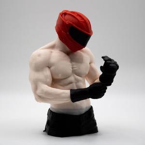 May include: A muscular torso figurine wearing a bright red helmet with a black visor and black gloves. The figure has a pale skin tone, defined chest and arm muscles, and is depicted adjusting the black gloves on its hands while facing downward.