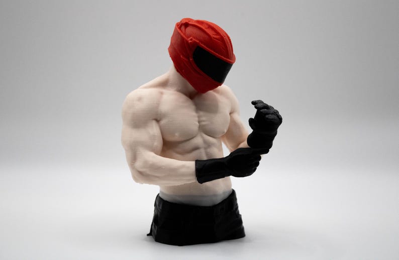 May include: A muscular torso figurine wearing a bright red helmet with a black visor and black gloves. The figure has a pale skin tone, defined chest and arm muscles, and is depicted adjusting the black gloves on its hands while facing downward.