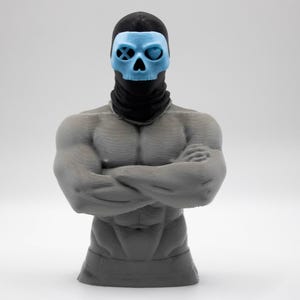 May include: A gray muscular torso figurine with arms crossed, wearing a black balaclava and a light blue skull mask. The mask features a circular eye socket with an X symbol on the right and a heart-shaped eye socket on the left side.