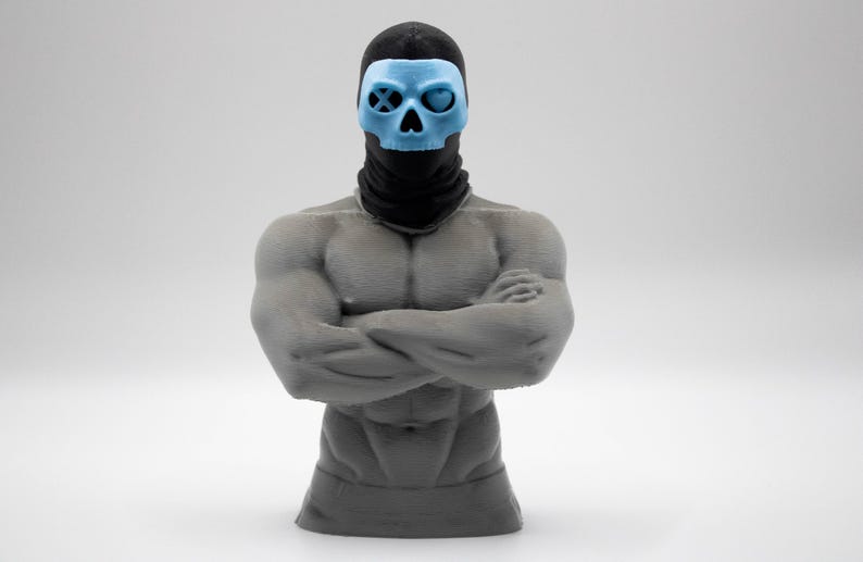 May include: A gray muscular torso figurine with arms crossed, wearing a black balaclava and a light blue skull mask. The mask features a circular eye socket with an X symbol on the right and a heart-shaped eye socket on the left side.