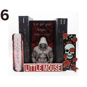 May include: A display featuring a hooded figure statue, books, and decorative elements. The books have titles like "Haunting Adeline" and "Cat got your tongue, little mouse?" A sign reads "Little Mouse". The color scheme is black, red, and white.