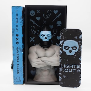 May include: A book titled "LIGHTS OUT" by Navessa Allen, with a blue cover. A black box with a skull-shaped figure and a matching bookmark. The box has light blue skull and medical symbol accents.