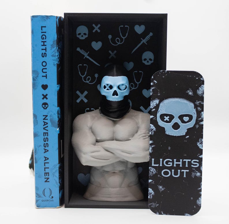 May include: A book titled "LIGHTS OUT" by Navessa Allen, with a blue cover. A black box with a skull-shaped figure and a matching bookmark. The box has light blue skull and medical symbol accents.