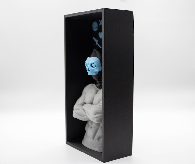 May include: A black rectangular shadow box contains a gray statue of a muscular figure with arms crossed. A light blue skull mask is on the figure's face. Light blue skull and crossbones, heart, and knife icons are on the back wall.