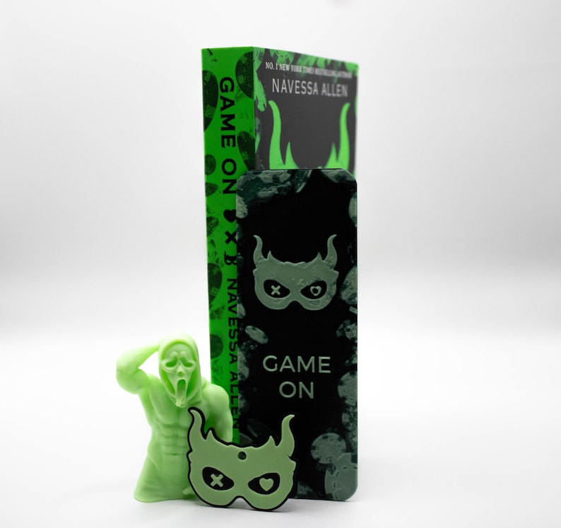 May include: A light green figurine of a screaming figure, a green mask-shaped charm, and a book with the title "Game On" and the author's name, Navessa Allen. The book and bookmark have a green and black design.