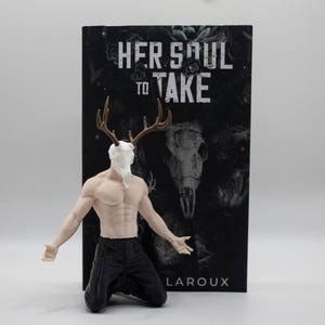 May include: A figurine of a muscular person kneeling, wearing black pants, with a white deer skull head and brown antlers. Behind the figurine is a book titled Her Soul to Take by Harley Laroux, featuring a dark cover with a skull and floral design.