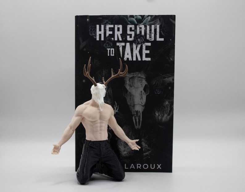 May include: A figurine of a muscular person kneeling, wearing black pants, with a white deer skull head and brown antlers. Behind the figurine is a book titled Her Soul to Take by Harley Laroux, featuring a dark cover with a skull and floral design.