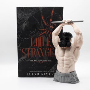 May include: A book titled Little Stranger by Leigh Rivers with a black cover featuring a red spider and web design, positioned next to a light-skinned muscular torso figurine wearing a black gas mask and grey shorts, lifting a grey barbell.