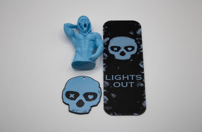 May include: A light blue Scream figure, a black bookmark with a skull design and the text "LIGHTS OUT", and a light blue skull-shaped tag with an X and a heart in the eyes. The items are on a white surface.