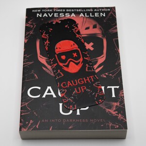 Pode incluir: Um livro intitulado "Caught Up" de Navessa Allen, com uma capa preta e vermelha e um marcador a condizer. O marcador apresenta um gr&aacute;fico vermelho de um capacete e as palavras "Caught Up". Um "Into Darkness Novel".