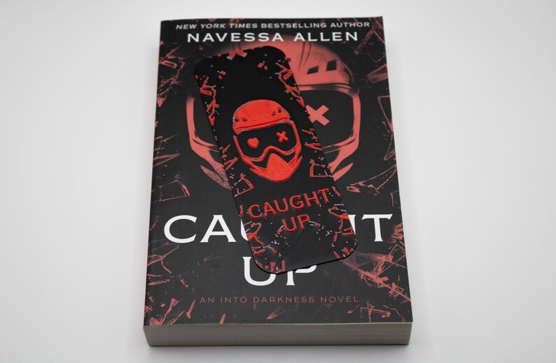 Pode incluir: Um livro intitulado "Caught Up" de Navessa Allen, com uma capa preta e vermelha e um marcador a condizer. O marcador apresenta um gr&aacute;fico vermelho de um capacete e as palavras "Caught Up". Um "Into Darkness Novel".