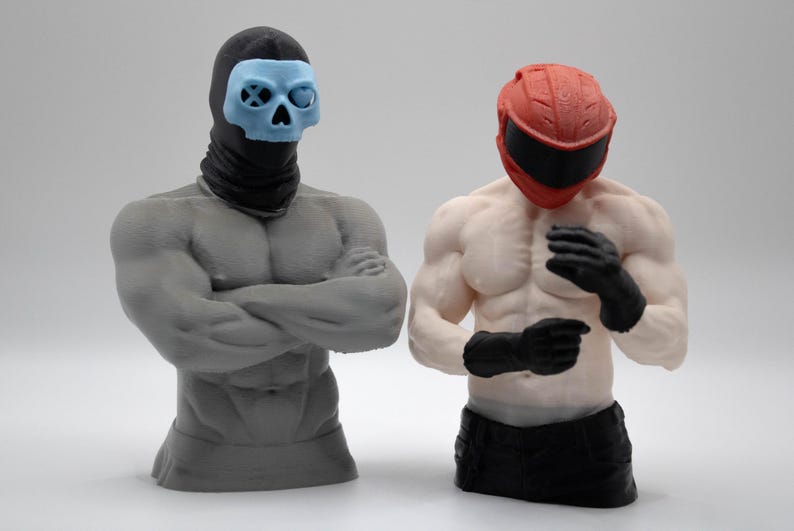 May include: Two muscular torso figurines. The left figure is gray with a black balaclava and a light blue skull mask. The right figure is skin-toned with a red motorcycle helmet and black gloves, wearing black pants. Both are standing upright.