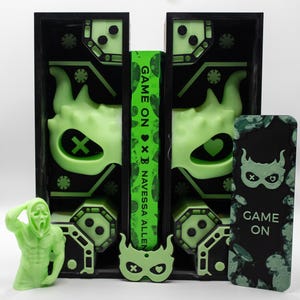 May include: A neon green and black display featuring a book with the title "Game On" and the author's name, Navessa Allen. The display includes a figurine, a mask, and a decorative panel with a mask design. The overall theme is a horror-themed book.