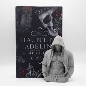 May include: A book titled Haunting Adeline by H.D. Carlton with a skull cover design placed next to a gray, 3D-printed torso figurine. The figurine depicts a muscular figure wearing a hooded sweatshirt with the hood pulled up over the head.