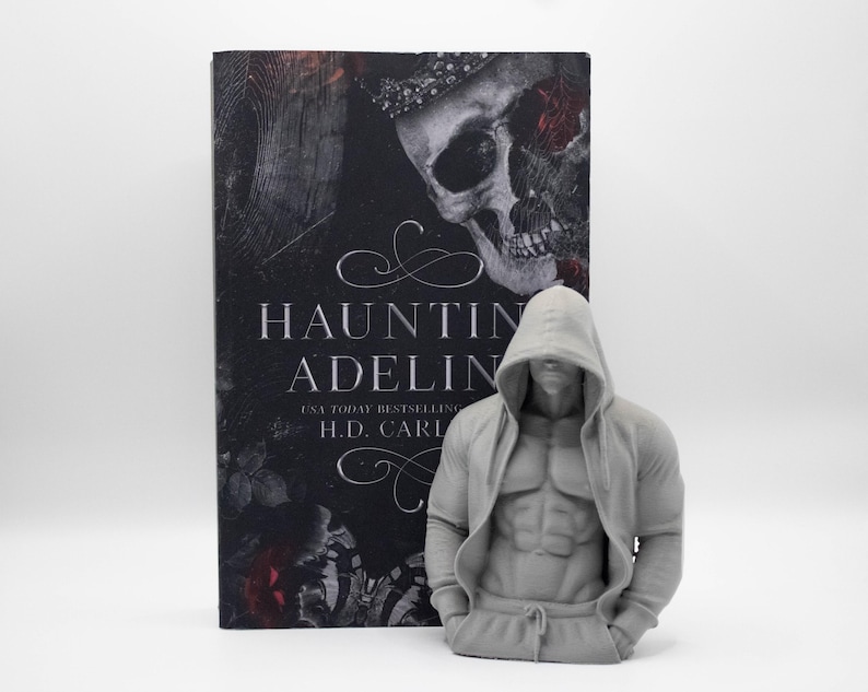 May include: A book titled Haunting Adeline by H.D. Carlton with a skull cover design placed next to a gray, 3D-printed torso figurine. The figurine depicts a muscular figure wearing a hooded sweatshirt with the hood pulled up over the head.