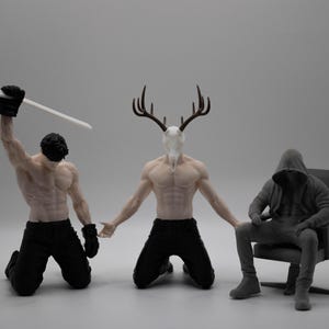 May include: Three figurines: one kneeling figure holding a white hockey stick, a central kneeling figure with a deer skull head and antlers, and a third figure in a hooded sweatshirt sitting on a chair, all featuring detailed muscular anatomy.
