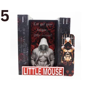 May include: A bookend set featuring a muscular figure in a hooded jacket, with the text "Cat got your tougur, little mouse?" and "LITTLE MOUSE". The set includes two books and a bookmark with a similar design. The books are titled "HAUNTING ADELINE" by H.D. Carlton.