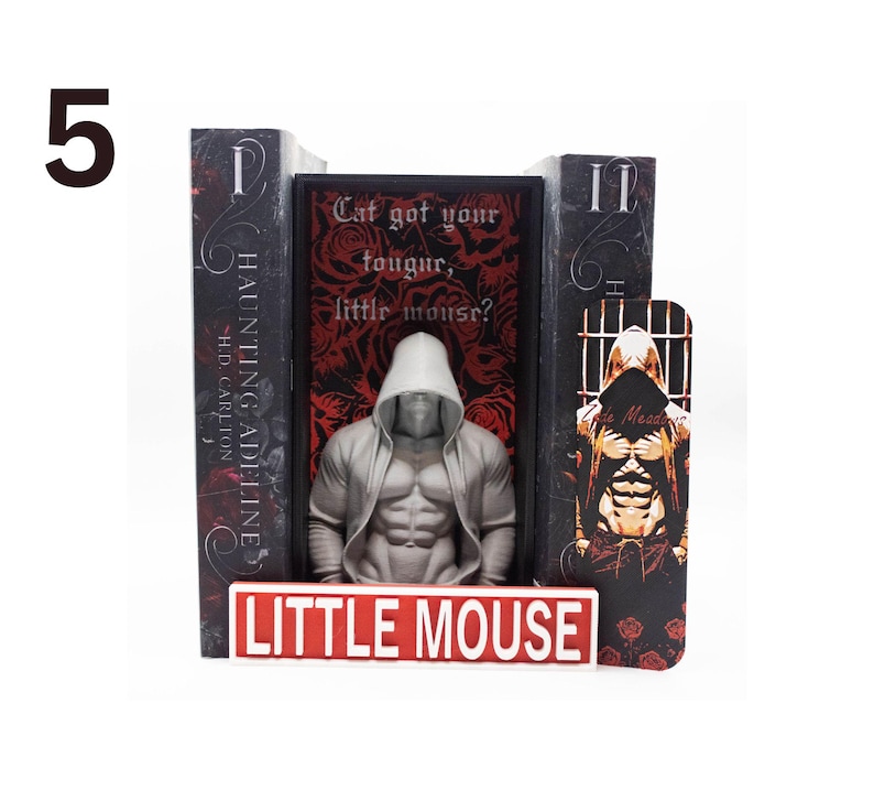 May include: A bookend set featuring a muscular figure in a hooded jacket, with the text "Cat got your tougur, little mouse?" and "LITTLE MOUSE". The set includes two books and a bookmark with a similar design. The books are titled "HAUNTING ADELINE" by H.D. Carlton.